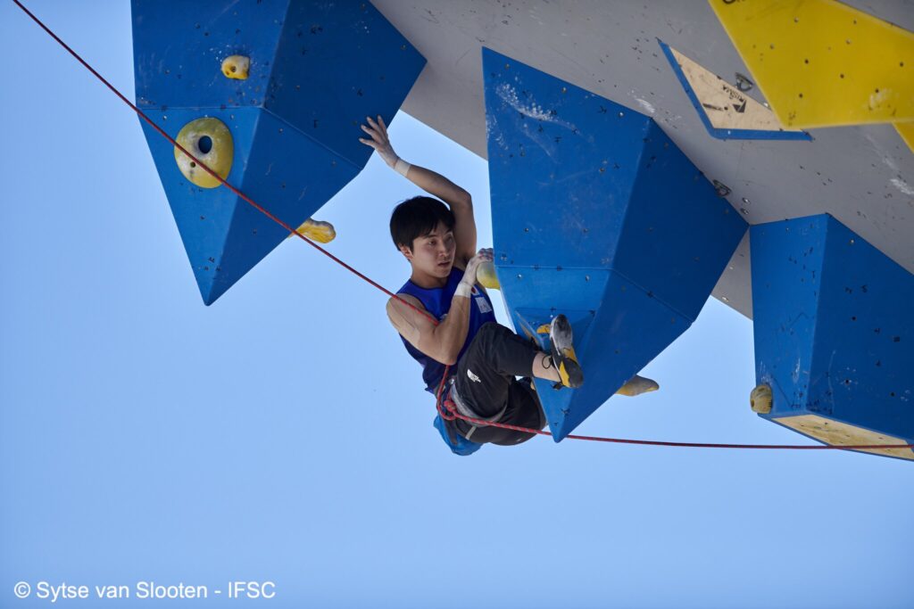 A climber is on a steep competition wall and is reaching for a huge volume overhead.