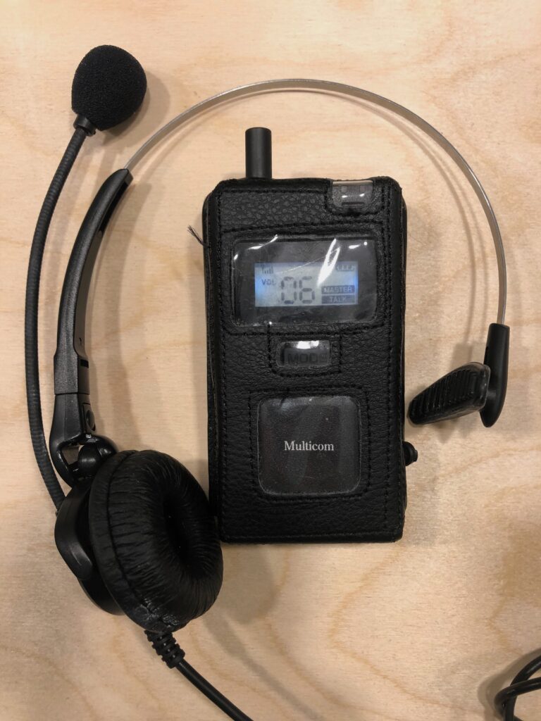 The swatcom handset and headset. It's a small black device in a case. The headset has a speaker on one ear and a microphone on a short boom.