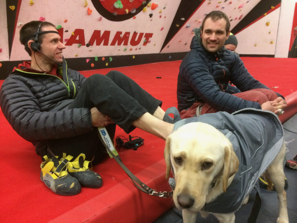 John sits in a climbing centre on boulder safety matting. He has his bare feet tucked under his guide dogs coat. Blind climber Jesse is smiling next to him. The dog isn't looking too happy.