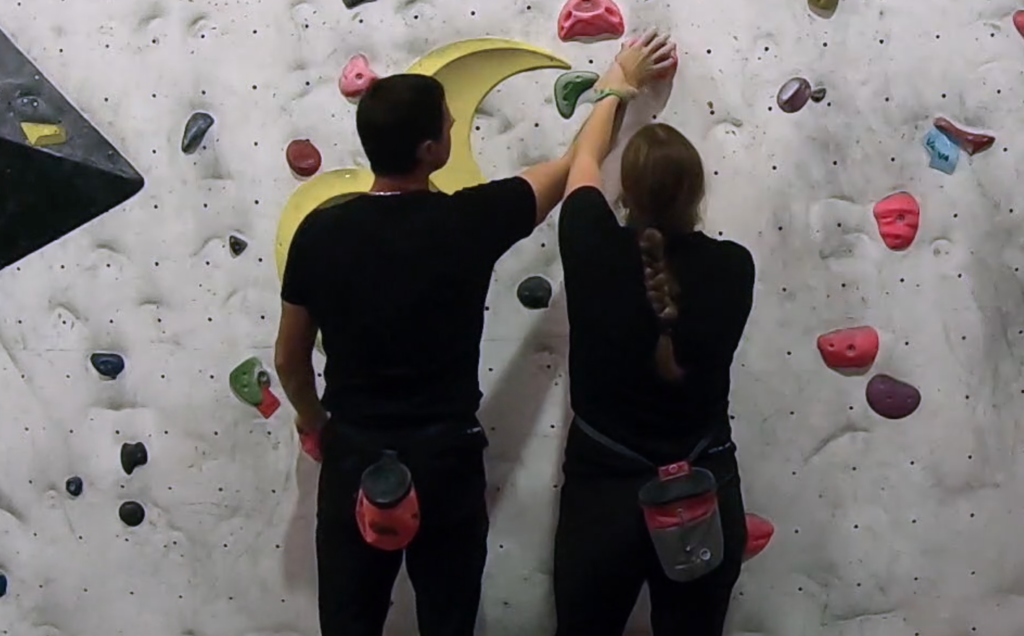 Stood in front of a climbing wall, Lauren guides John's hands to a climbing hold. 
