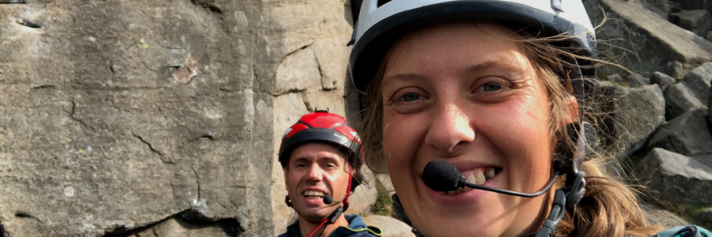 Lauren and John aree stood in front of a steep rock face wearing helmets and a microphone.