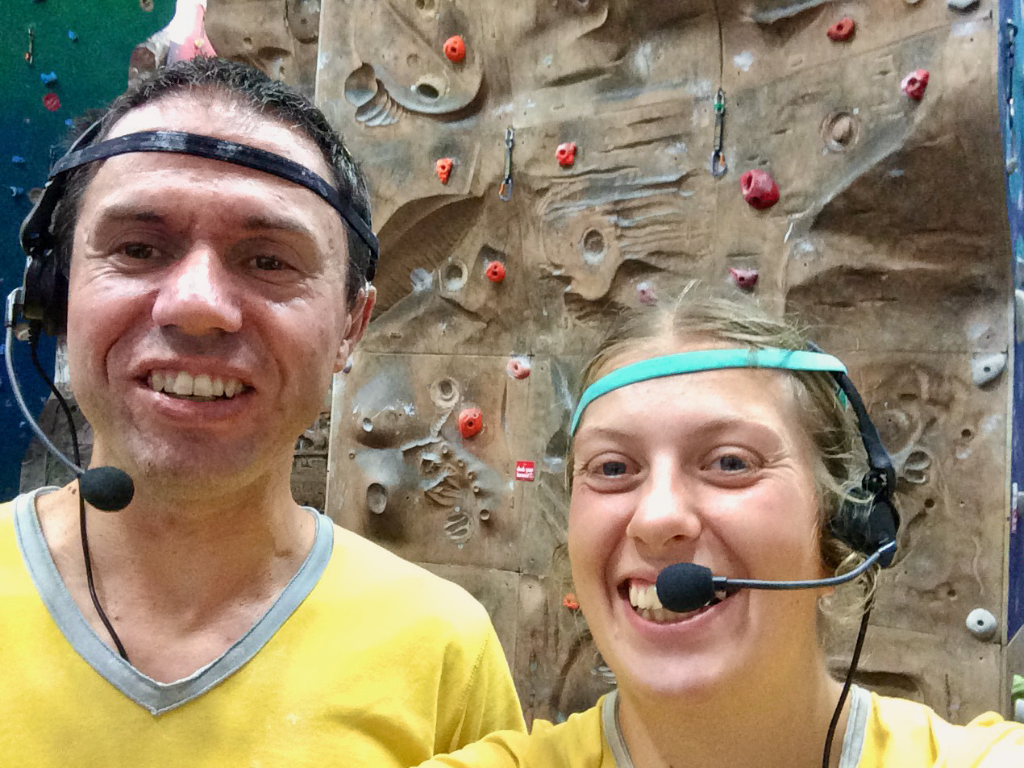 Lauren and John each wear a headset with a microphone at an indoor climbing wall